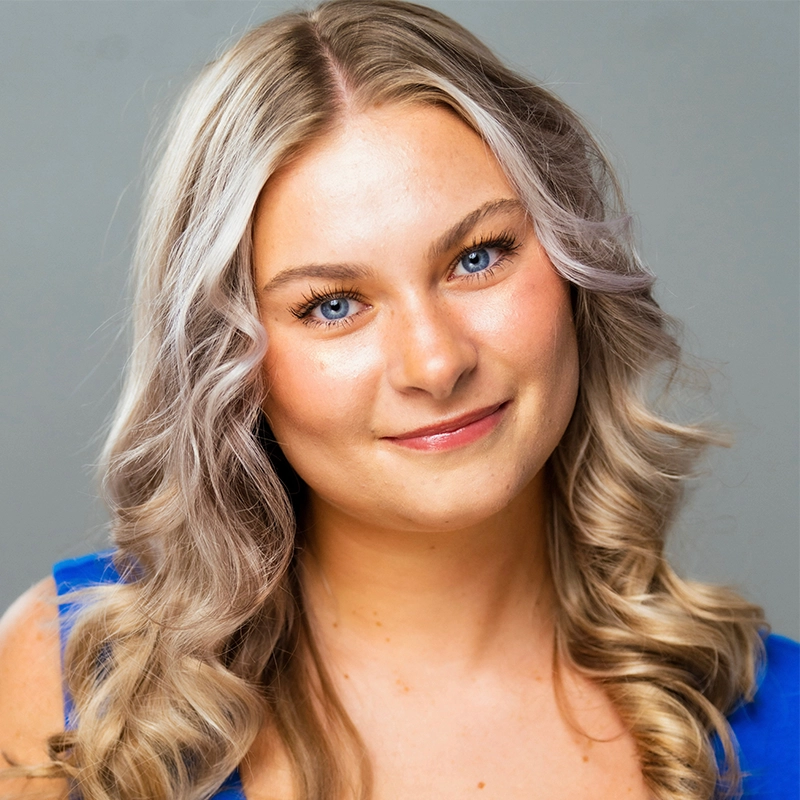 A young woman with long, wavy blonde hair smiles gently at the camera. She has fair skin, blue eyes, and is wearing a bright blue top. The background is plain and gray, drawing all attention to Saylor Tucker’s warm and inviting expression.