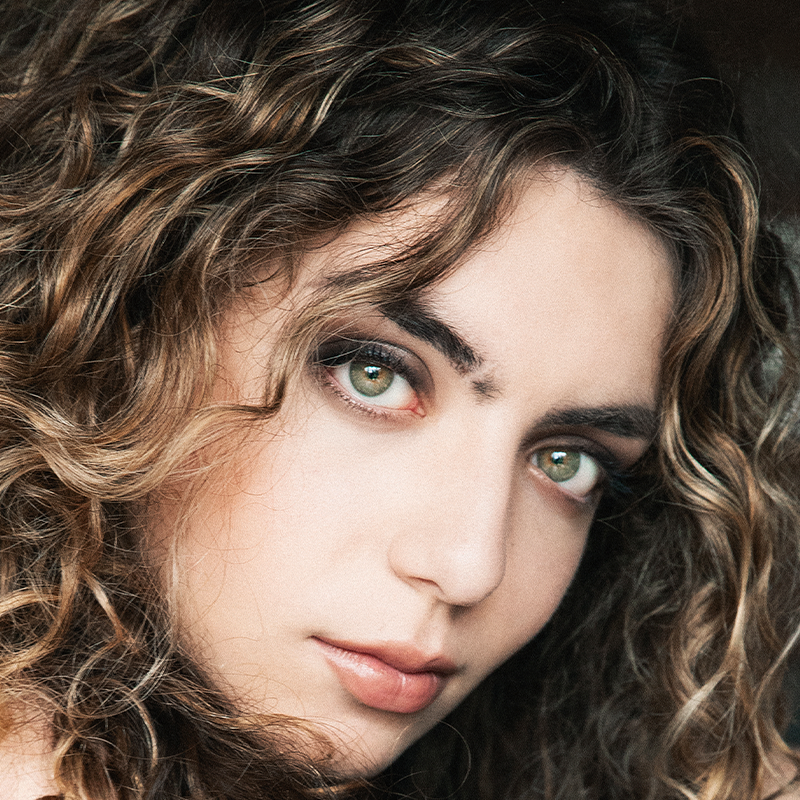 A young person with light skin and curly, light brown hair gazes intently at the camera. They have green eyes, strong brows, and natural makeup. The background is out of focus, highlighting their face and hair. The lighting is soft, creating a gentle mood that perfectly captures Elle Gurevich’s expressive features.