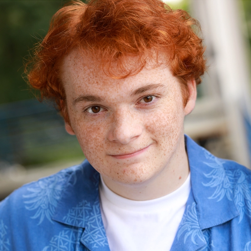 A young person with curly red hair and freckles smiles softly at the camera. They are wearing a blue patterned shirt over a white T-shirt. The background is blurred and appears to be outdoors, capturing a candid moment reminiscent of Millie Yocum Surfac’s vibrant spirit.