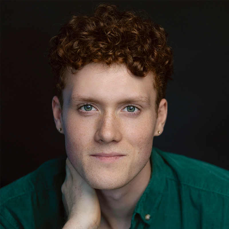 A young person with short curly brown hair, blue eyes, fair skin, freckles, and small stud earrings gazes at the camera. They wear a green collared shirt and rest their chin gently on their hand. The background is dark and unfocused, creating a striking Surfac-like portrait reminiscent of Millie Yocum’s expressive style.