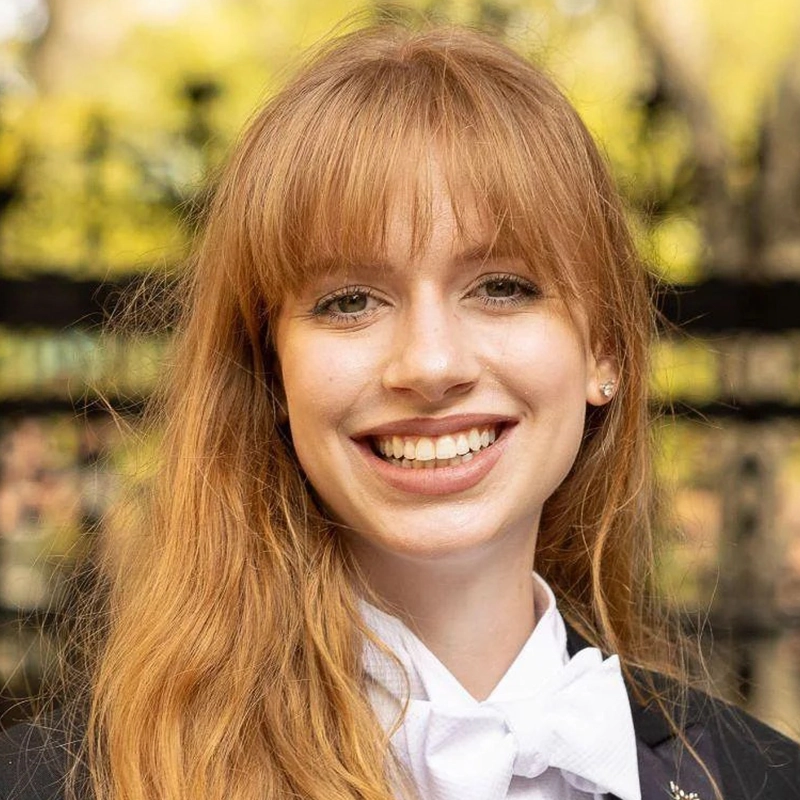A young woman with long, straight red hair and bangs smiles broadly. She wears a black jacket and a white shirt with a bow tie, resembling the spirited style of Millie Yocum. The blurred background shows greenery and a dark fence, giving the photo an effortless Surfac aesthetic.