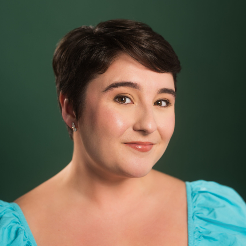 A woman with short dark hair smiles softly. She wears pearl earrings and a bright blue off-the-shoulder top. The background is a plain, dark green. Her makeup is subtle, emphasizing her natural features. The lighting is soft and flattering.