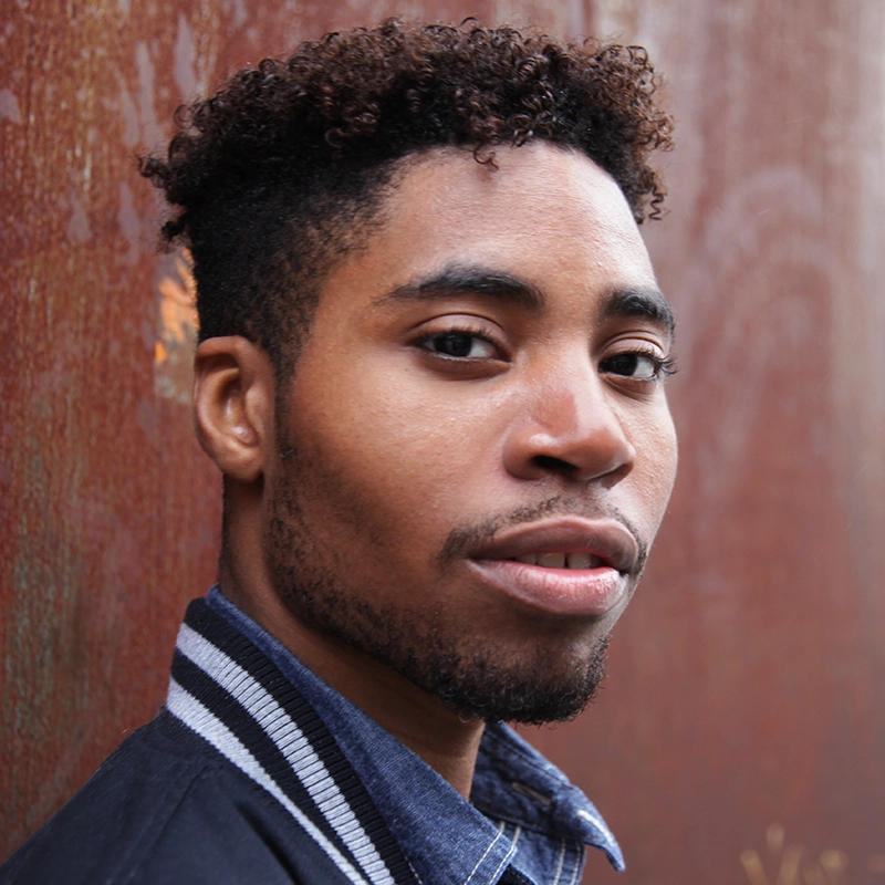 A young man with short curly hair and a trimmed beard looks at the camera with a slight smile. He wears a striped-collar jacket over a denim shirt and stands against a textured, rusty brown wall in natural light.