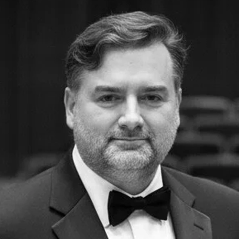 A man with short, wavy hair and a beard wears a black tuxedo and bowtie. He faces the camera with a neutral expression. The background is blurred and dark, suggesting an indoor setting. The image is in black and white.