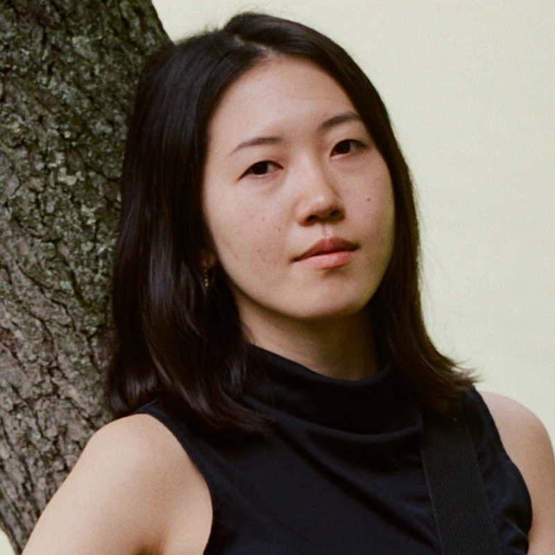 A young woman, Juliann Frances, with straight, shoulder-length black hair stands outdoors next to a tree. She wears a sleeveless black top and looks at the camera with a neutral expression, the softly blurred background emphasizing her face and the tree trunk.
