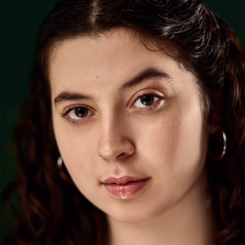 A young woman, Jayline Ross, with light skin, brown eyes, and long dark curly hair is shown in close-up. She wears silver hoop earrings and a neutral expression. The softly blurred dark green background emphasizes her face.