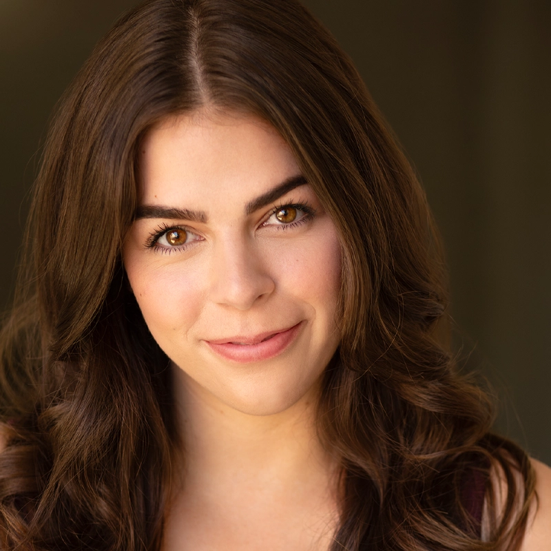 A young woman with long, wavy brown hair smiles softly. She has light skin, brown eyes, and softly arched brows. The background is dark and blurred, drawing focus to her face. She is wearing a sleeveless top and natural makeup.
