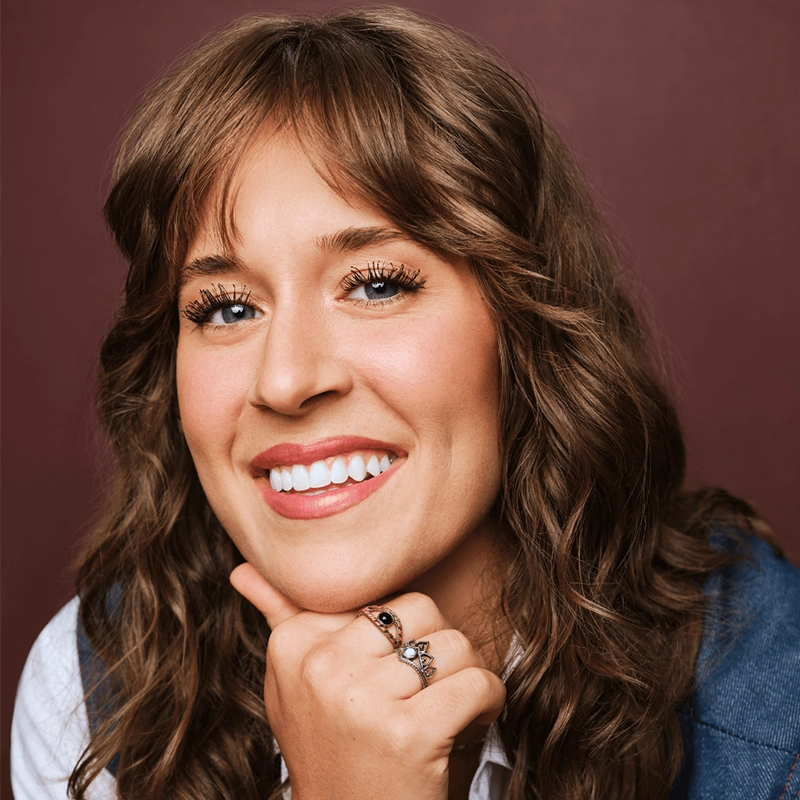 A smiling woman with wavy brown hair and blue eyes poses in front of a maroon background. Channeling Aaron Valentine’s warm style, she rests her chin on her hand, displaying rings, and wears a white shirt with a blue vest.