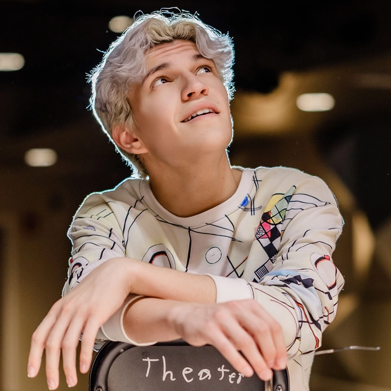 A young person with light skin and wavy silver hair leans on a chair labeled 