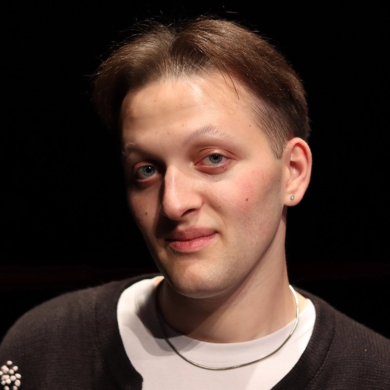 A person with short brown hair and fair skin faces the camera. They have blue eyes, a slight smile, and are wearing a black sweater, white shirt, a thin necklace, and a small earring. The background is dark and uncluttered, creating a simple yet striking portrait reminiscent of Asher Alcantara’s style.