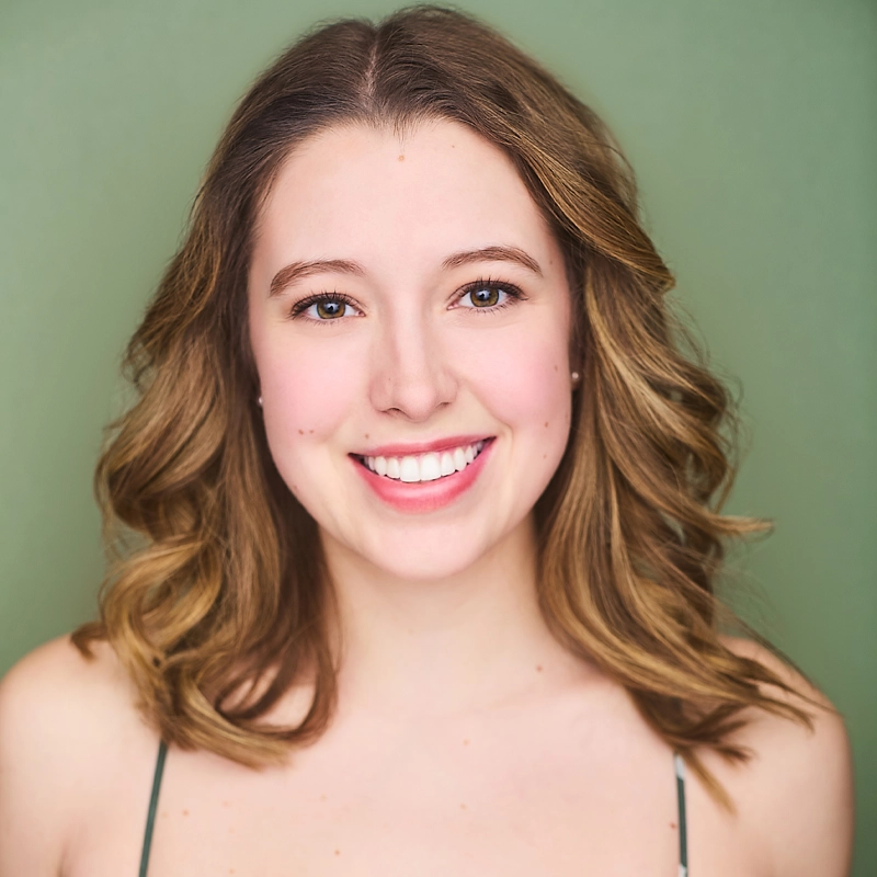 Young woman, Ellie Falanga, with wavy light brown hair, wearing a sleeveless top, smiles warmly at the camera against a plain green background. Her skin is fair, and she has natural makeup with a focus on her eyes and bright smile.