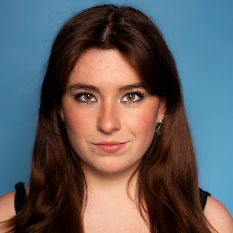 A young woman with long brown hair, fair skin, and hazel eyes looks directly at the camera. She wears subtle makeup, a black sleeveless top, and small hoop earrings. The background is a solid light blue, creating a clean and modern portrait reminiscent of Kailyn Fetterman's signature style.