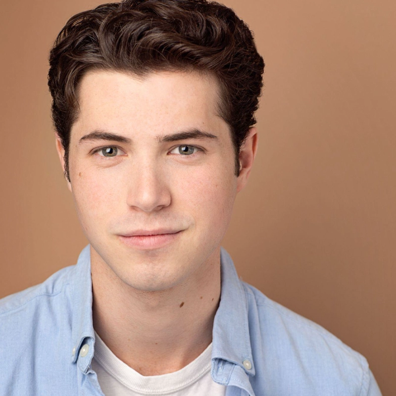 A young man with short, wavy dark hair and light skin is wearing a light blue button-up shirt over a white t-shirt. With a soft smile, Broderik gazes directly at the camera against a plain, warm beige background.