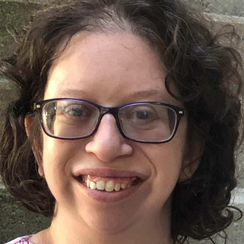 Rachel Flamm, a person with short, curly brown hair and glasses, smiles at the camera. They have light skin and are outdoors, with a blurred stone wall in the background. The photo is a close-up, and the lighting is natural.