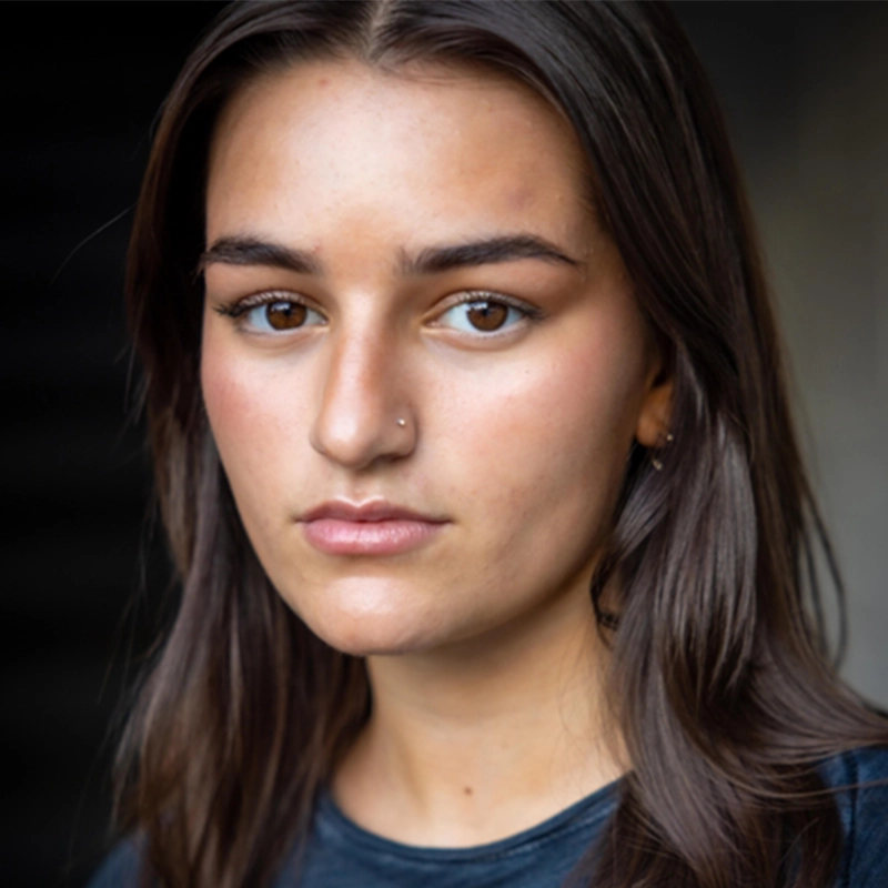 A young woman with straight dark brown hair and brown eyes looks into the camera with a neutral expression. She has a small nose piercing and wears a dark shirt. The background is blurred, highlighting her face as the main focus—a subtle confidence that perfectly captures the 