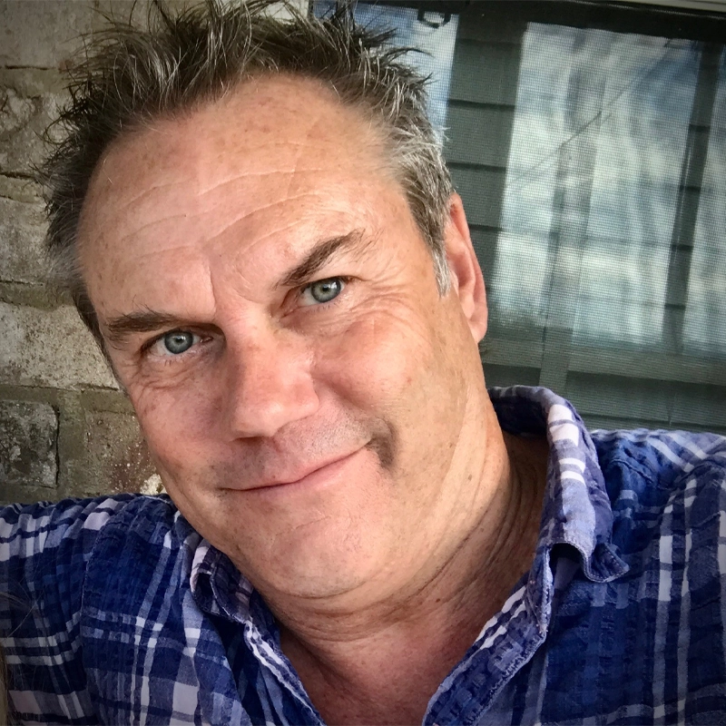 A middle-aged man with short, spiked gray hair and blue eyes smiles at the camera. Glen Fretwell wears a blue plaid shirt and is outdoors in front of a stone wall and window with a screen. Sunlight highlights his face.