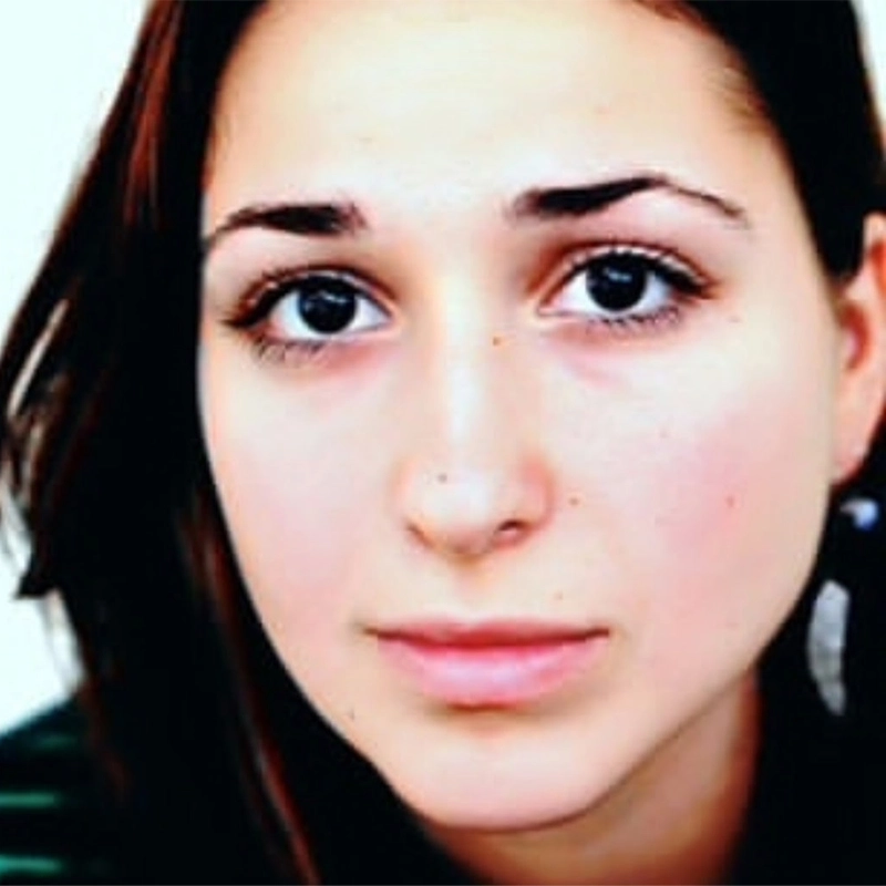 A young woman with long dark hair and brown eyes looks directly at the camera with a neutral, slightly serious expression. The background is plain and light-colored. Gabby Sherba wears a striped dark top and a single visible earring.