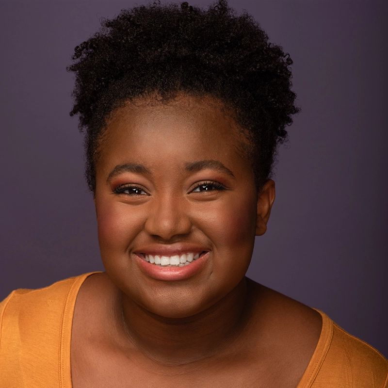 Gabrielle Farley, a young woman with curly hair, smiles broadly. She wears a mustard yellow top and subtle makeup. The background is a plain, solid purple. The lighting highlights her joyful expression and glowing skin.