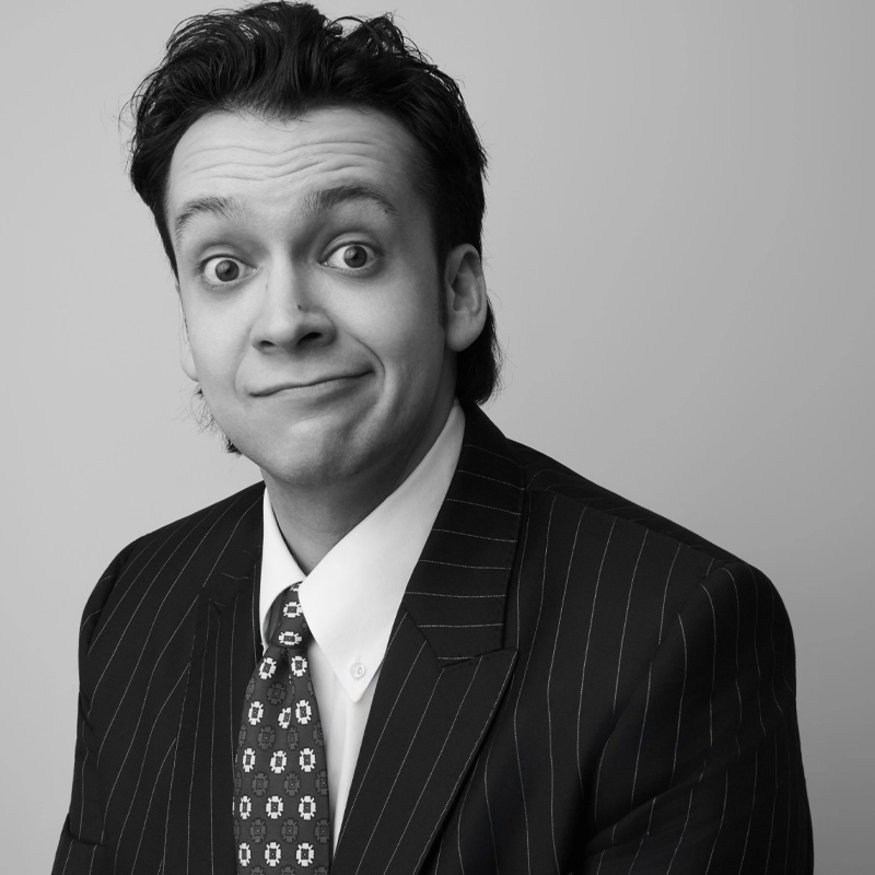 A man in a pinstripe suit and patterned tie, with dark hair, looks at the camera with raised eyebrows and a slight, crooked smile. The background is plain and grey, giving the black-and-white portrait of Fernando Zermeño Garavito a formal yet humorous feel.