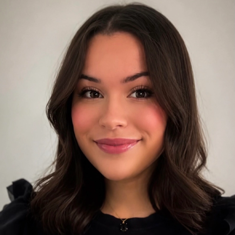 Victoria Grabois, a young woman with wavy, shoulder-length brown hair, smiles gently. She has smooth skin, subtle makeup with pink blush and lipstick, and is wearing a black top. The background is plain and light-colored.