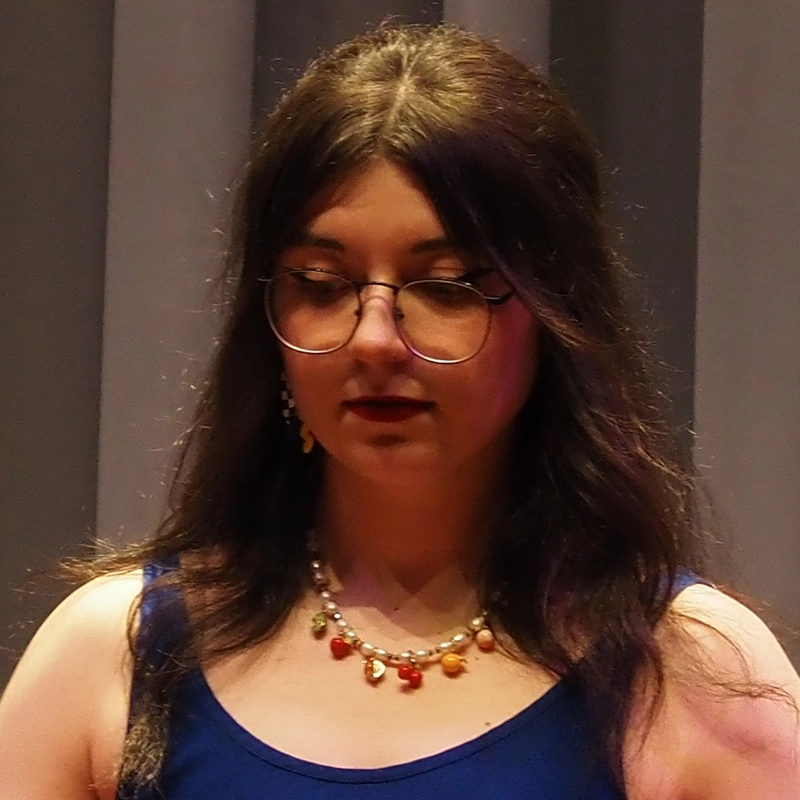 Juliann Frances, with long dark hair and round glasses, looks down. She wears a blue sleeveless top, a colorful beaded necklace, and drop earrings. The background features vertical gray panels with soft lighting.