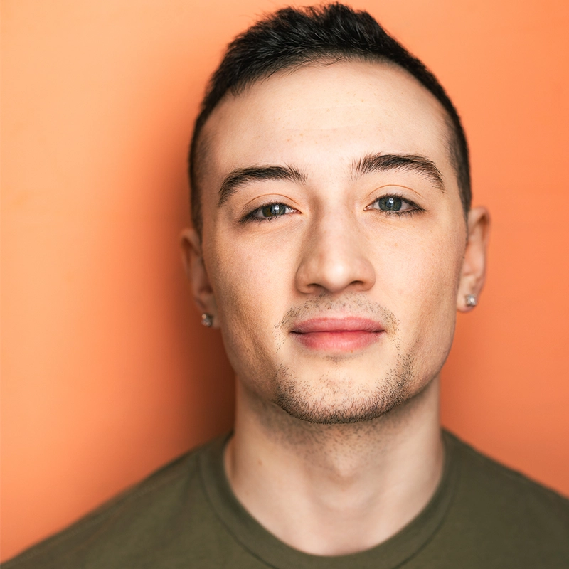 A young person with short dark hair, light skin, stud earrings, and slight facial hair looks directly at the camera, lightly smiling. They wear an olive green shirt and stand against a solid orange background—an expressive portrait reminiscent of Michelle Zink-Muñoz's candid style.