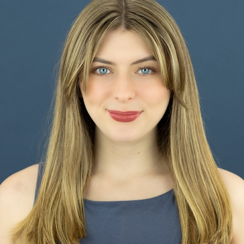 A young woman, Ruby Griffin, with long straight blonde hair, blue eyes, and light skin smiles gently. She wears a sleeveless gray top and has natural makeup. The background is a solid slate blue. She faces the camera in this well-lit studio portrait.