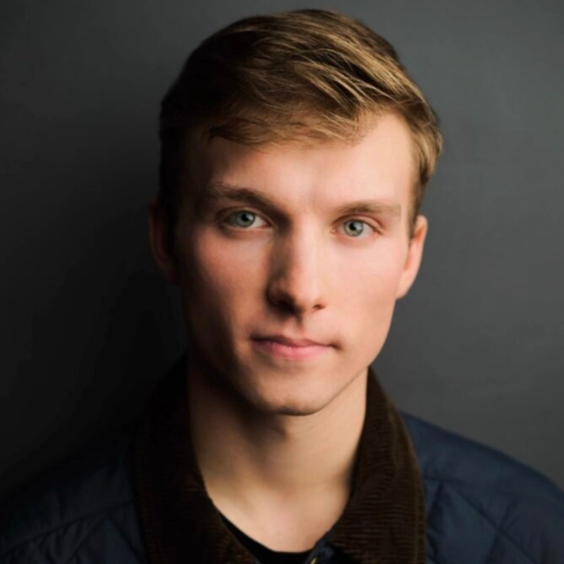A young man, Asher Alcantara, with light brown hair and green eyes looks directly at the camera, expression neutral. He wears a dark jacket over a shirt. The background is plain and gray, with soft lighting highlighting his face.