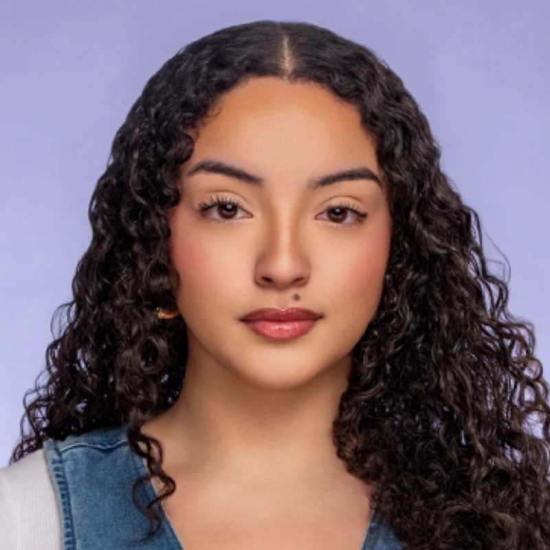A young person with long, dark curly hair and glowing skin stares directly ahead. They wear a denim top over a white shirt. The background is a plain, soft lavender color. Their expression is calm and neutral.