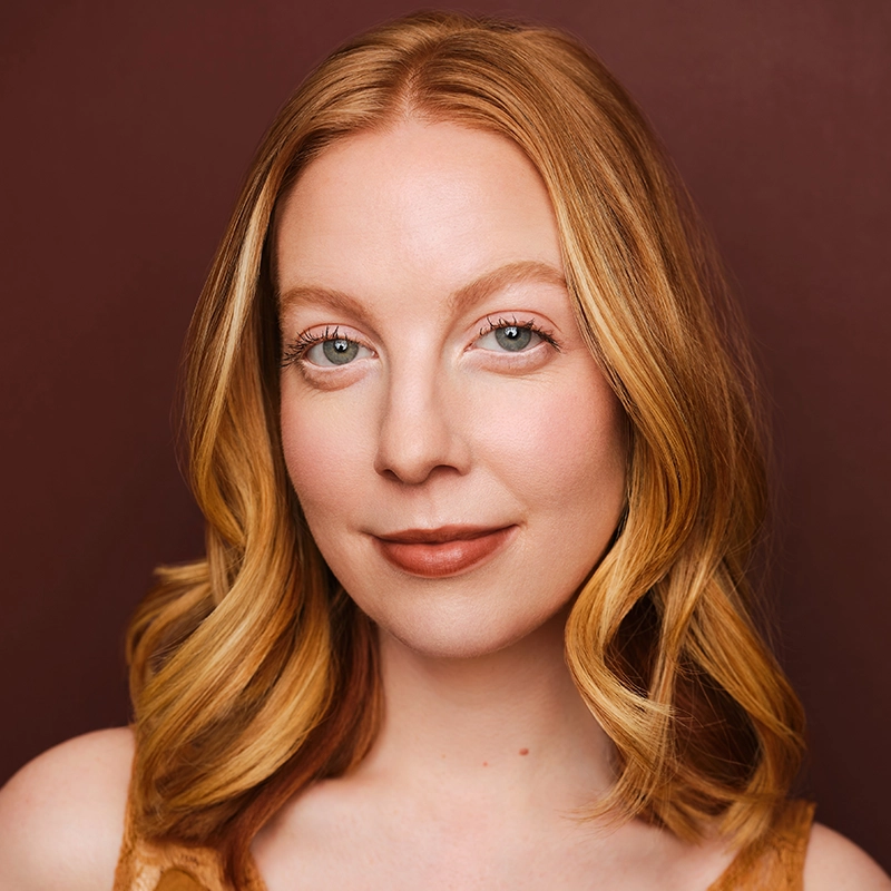 A woman with fair skin and shoulder-length wavy red hair smiles softly. She has blue eyes, natural makeup, and wears a light brown top. The background is a solid dark brown color. The lighting is soft, highlighting her facial features.