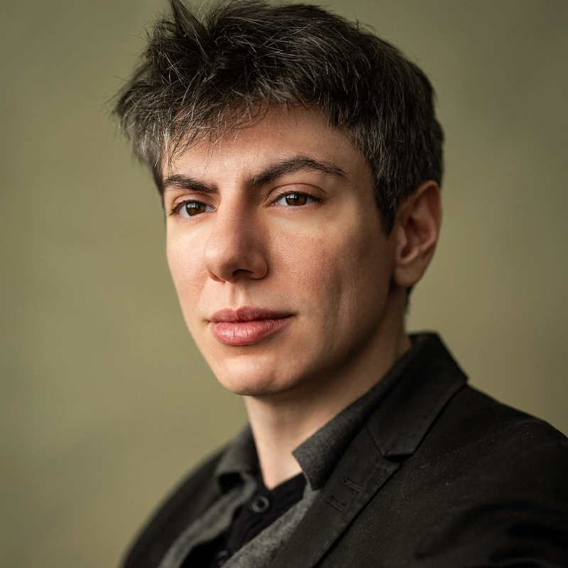 Charlie Hano, with short, tousled dark hair and fair skin, wears a dark blazer over a collared shirt. They face the camera with a soft, confident expression against a plain, warm-toned background as gentle lighting highlights their features.