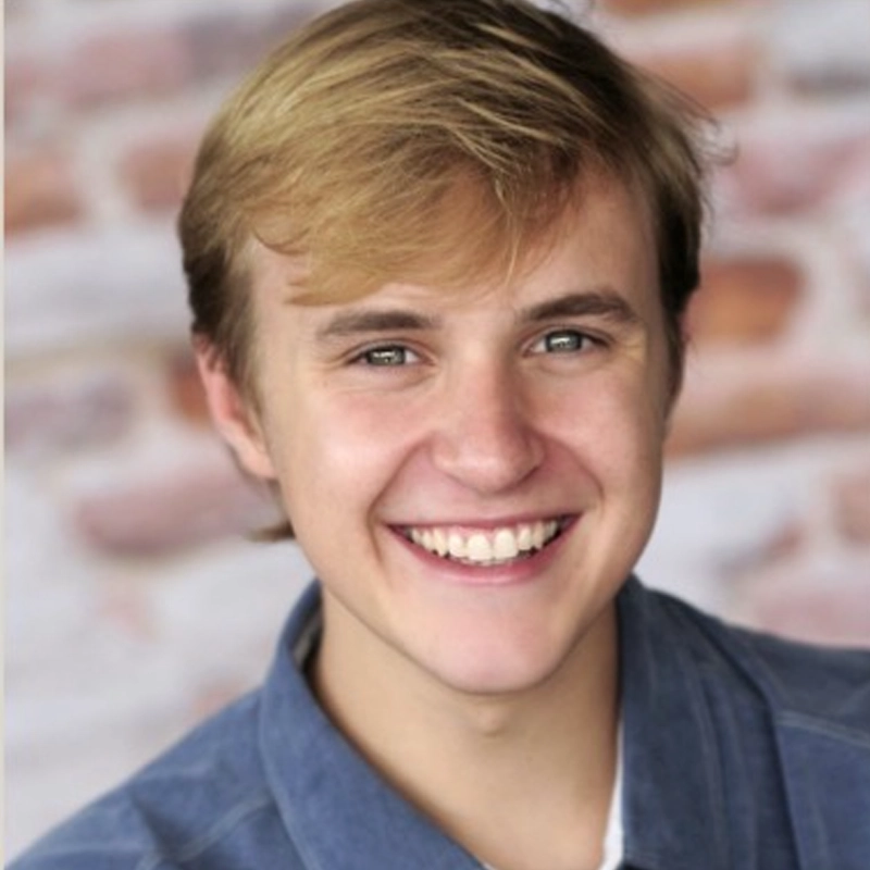 Carter Harvey, a young person with light brown hair and blue eyes, smiles at the camera in a blue collared shirt. The softly blurred background features tones of white, pink, and orange, resembling a brick wall with bright natural lighting.