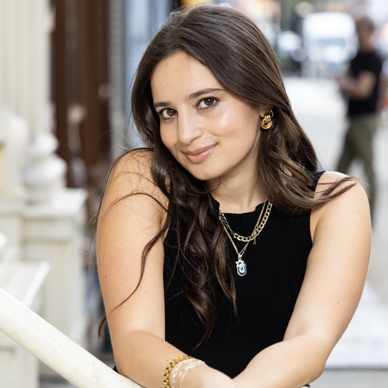 Chloe Hechter, a woman with long brown hair, wearing a black sleeveless top, gold jewelry, and a blue pendant necklace, leans on a white railing and smiles at the camera. The background shows a blurred city street scene with buildings and people.