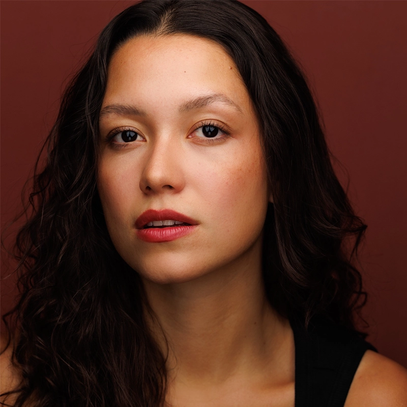 A young woman with long, wavy dark hair and light skin gazes confidently at the camera. She wears subtle makeup with red lipstick and a black top. The background is a solid burgundy color, creating a warm, focused portrait.