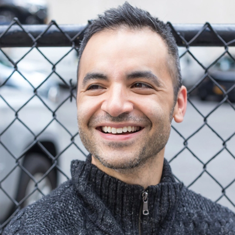 A man with short dark hair and stubble smiles, showing teeth. Rick Hip-Flores wears a dark zip-up sweater and stands in front of a chain-link fence with parked cars and a blurred building in the background. The photo is taken outdoors in daylight.