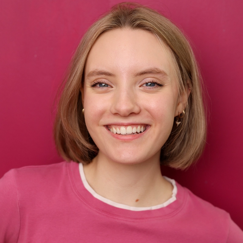 A young woman with light skin and short blonde hair smiles warmly. She wears gold hoop earrings and a pink top with a white collar. The background is a solid deep pink color. Her head is slightly tilted, and she looks directly at the camera.