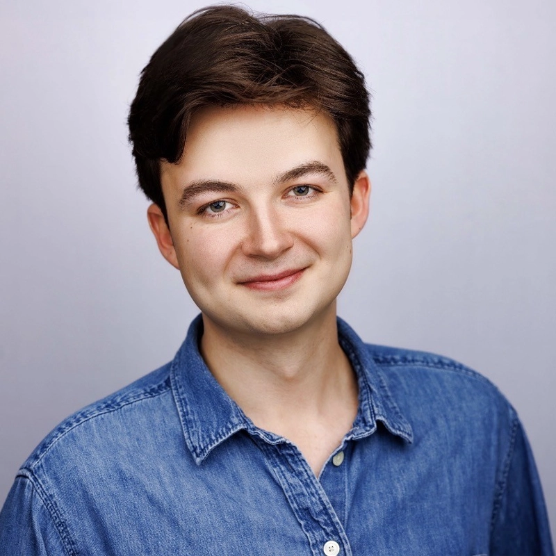 Jack Humphrey, a young man with fair skin, short brown hair, and brown eyes, smiles gently at the camera. He is wearing a medium blue denim shirt against a white background with soft, even lighting.