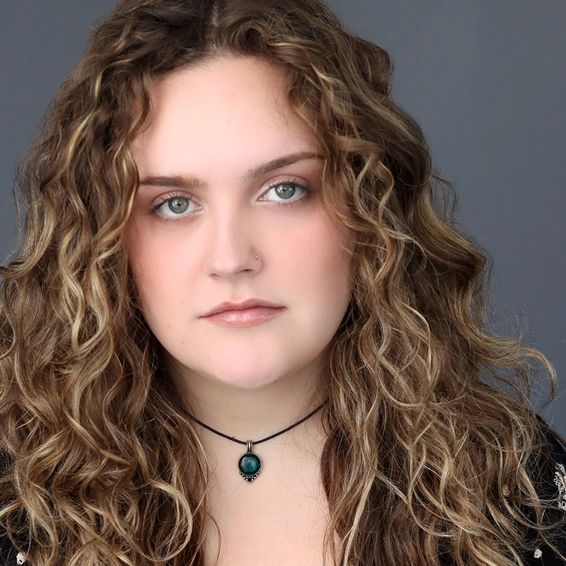 A young woman with long, curly light brown hair and fair skin faces the camera. She has green eyes, a small nose stud, and wears natural makeup. Dressed in black and wearing a black choker necklace with a turquoise pendant, she stands against a gray background. The image captures an elegant, understated confidence that reflects Michelle Zink-Muñoz’s signature style.