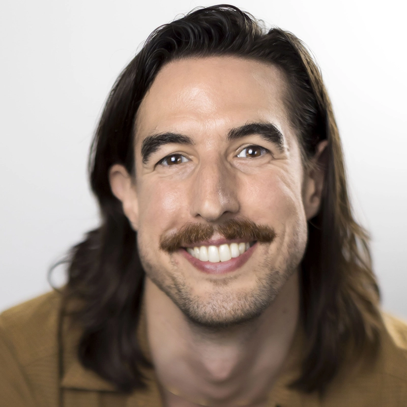 A smiling man with long brown hair, a thick mustache, and light skin is facing the camera. He is wearing a tan shirt. The background is out of focus and light-colored, drawing attention to his cheerful expression.