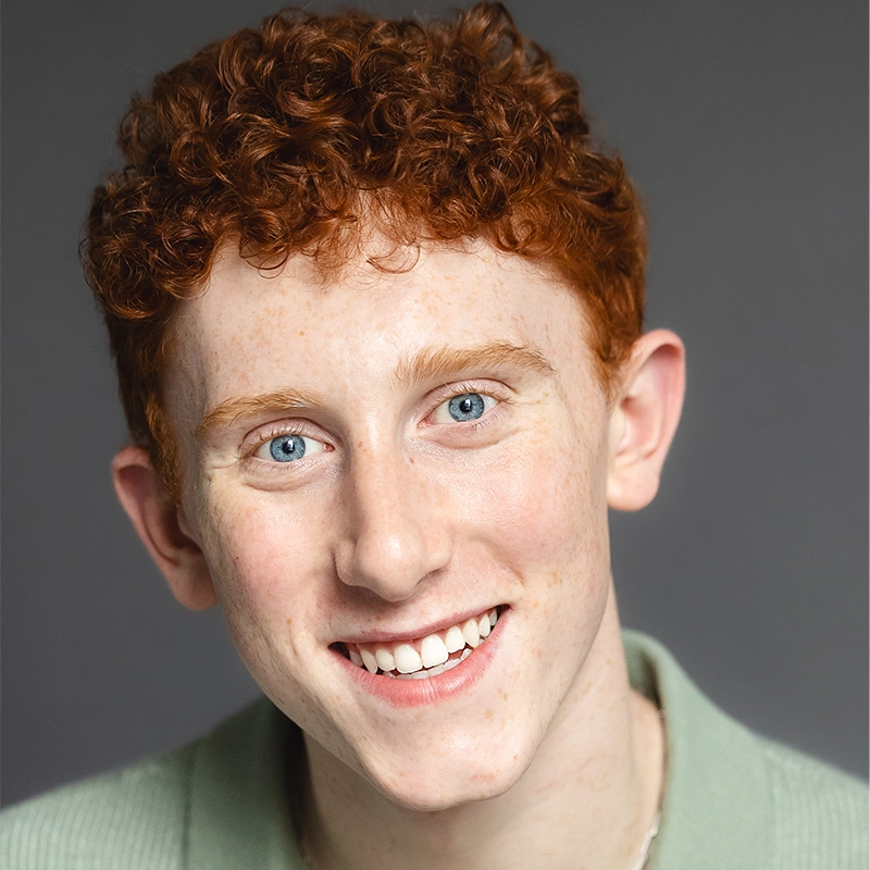 Young person with curly red hair, fair skin, and blue eyes smiles at the camera. They wear a light green collared shirt and are posed against a plain gray background. The soft lighting highlights their friendly expression, giving Millie Yocum a warm and approachable appearance in this Surfac portrait.