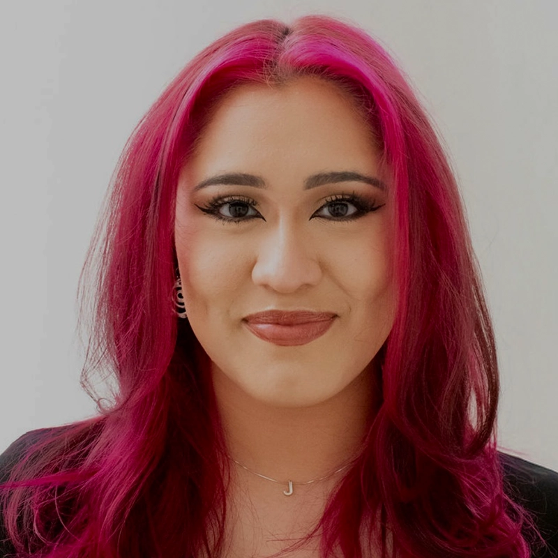 A person with long, vivid magenta hair and soft waves smiles gently. They have defined eyebrows, bold eye makeup, and wear nude lipstick. A silver necklace with a “J” pendant hints at their name—Jayline Ross. The background is plain and light-colored.
