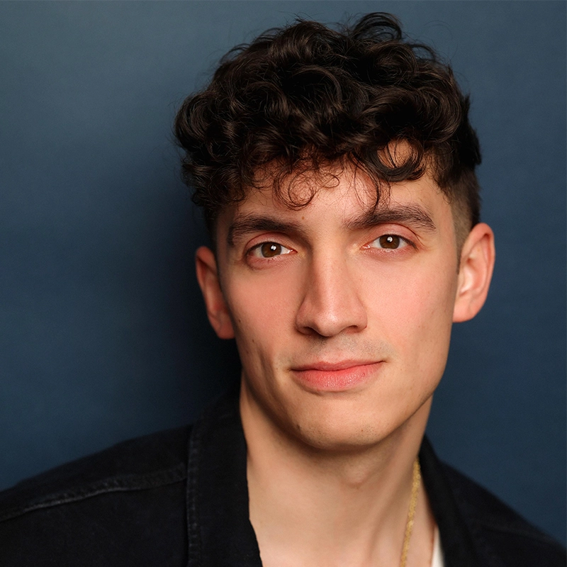 Young man with short, curly dark hair and fair skin poses against a plain dark blue background. He wears a black jacket over a light shirt and a thin gold chain. With a relaxed expression, James Olivas looks directly at the camera, offering a slight smile.