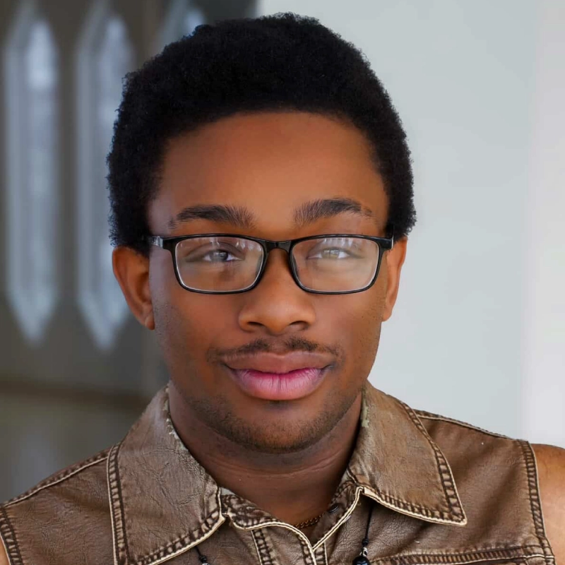 Jayline Ross, a young person with short curly hair and glasses, looks at the camera with a slight smile. They wear a brown leather jacket. The background is softly blurred, with light streaming through windows.