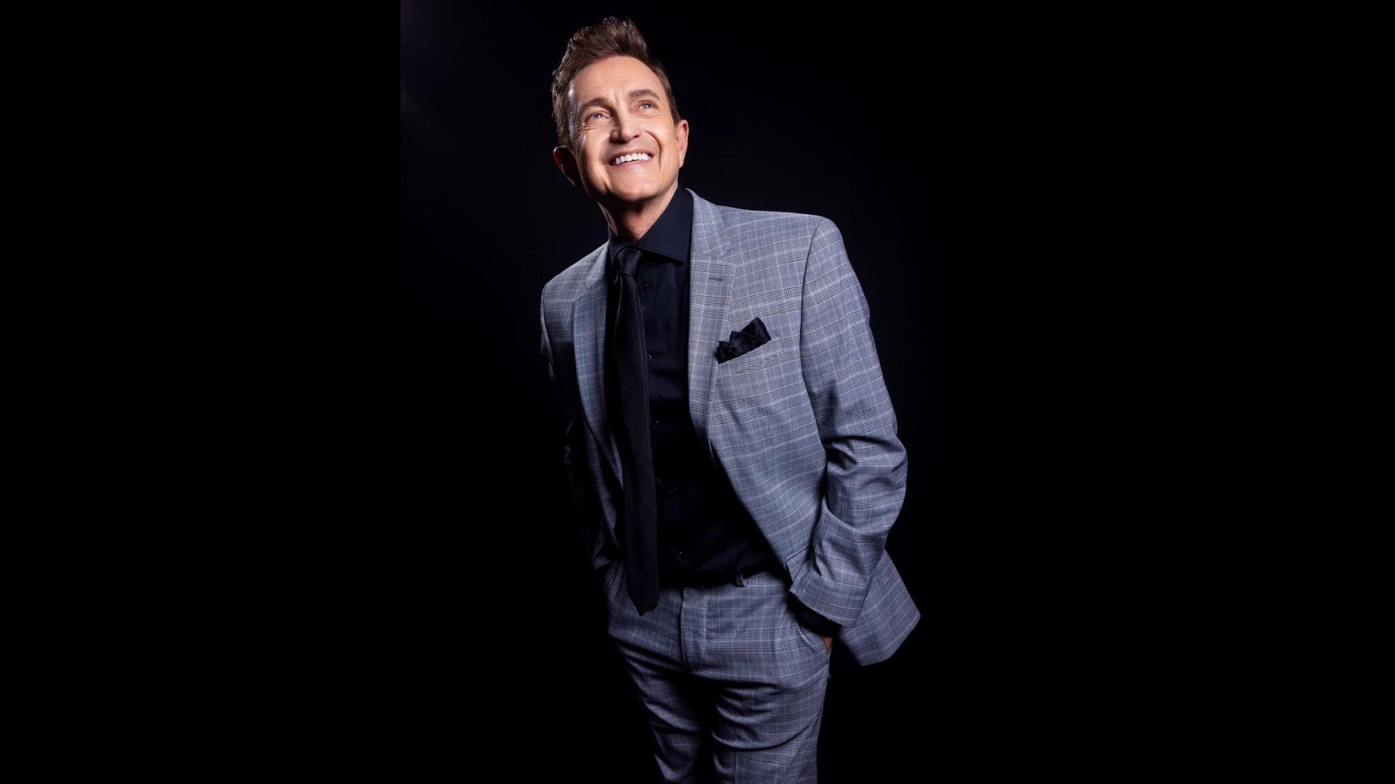 A man stands against a black background, smiling and looking upward. He wears a light gray plaid suit with a black shirt and tie, plus a black pocket square. With his left hand in his pocket, he appears relaxed and happy, as if channeling the smooth charm of Jeff Harnar performing 