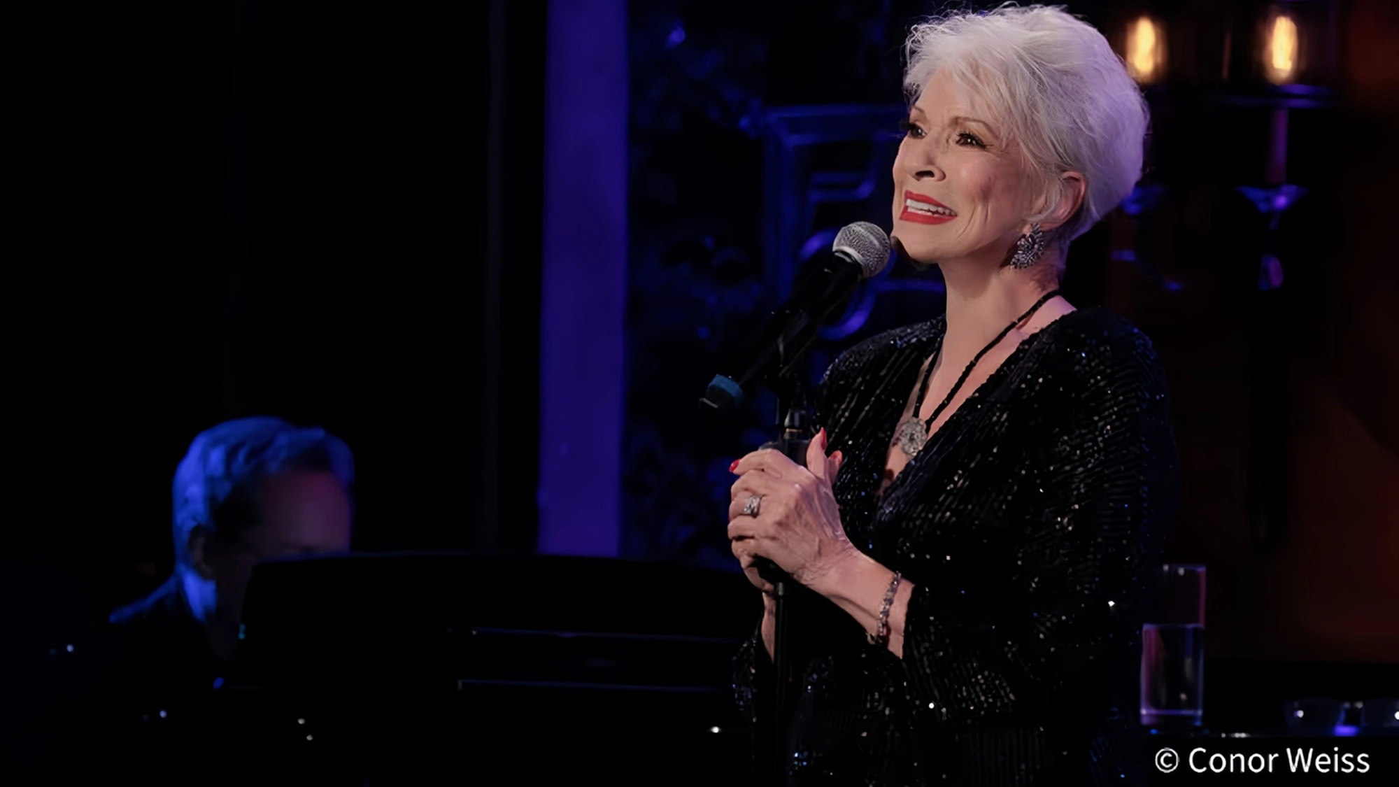 An older woman with short white hair, wearing a sparkly black outfit and red lipstick, sings into a microphone on stage. Reminiscent of Jennifer Bassey—best known for her roles in soaps—she smiles warmly, hands clasped around the mic. A pianist is blurred in the background. Stage lighting is blue and purple. © Conor Weiss.