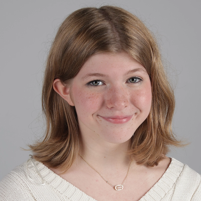 A young person with shoulder-length light brown hair and freckles smiles gently. They wear a cream knit sweater and a gold necklace with an oval pendant. The background is plain gray, highlighting Jenny Walker's calm and approachable presence.