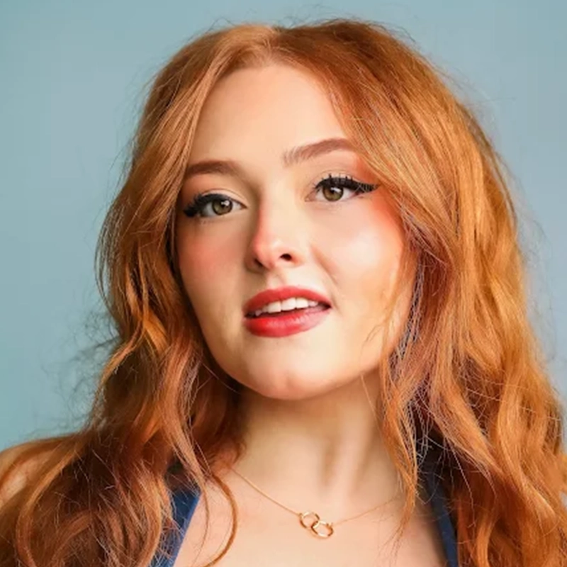 A young woman with long, wavy red hair poses in front of a pale blue background. She has fair skin, winged eyeliner, subtle blush, and red lipstick. Megan Ludwig wears a gold necklace with two linked rings and a blue top, looking confidently at the camera.