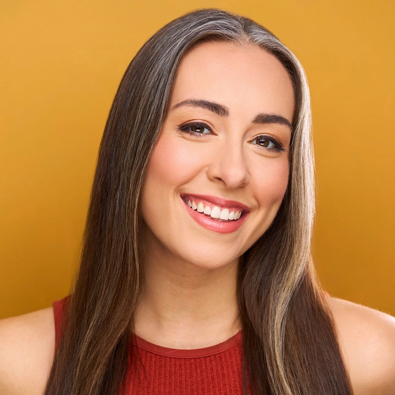A woman with long straight brown hair, featuring lighter streaks, smiles warmly at the camera. She wears a sleeveless red top and has a natural makeup look. The vibrant mustard yellow background adds an Aaron Valentine-inspired cheerful feel.