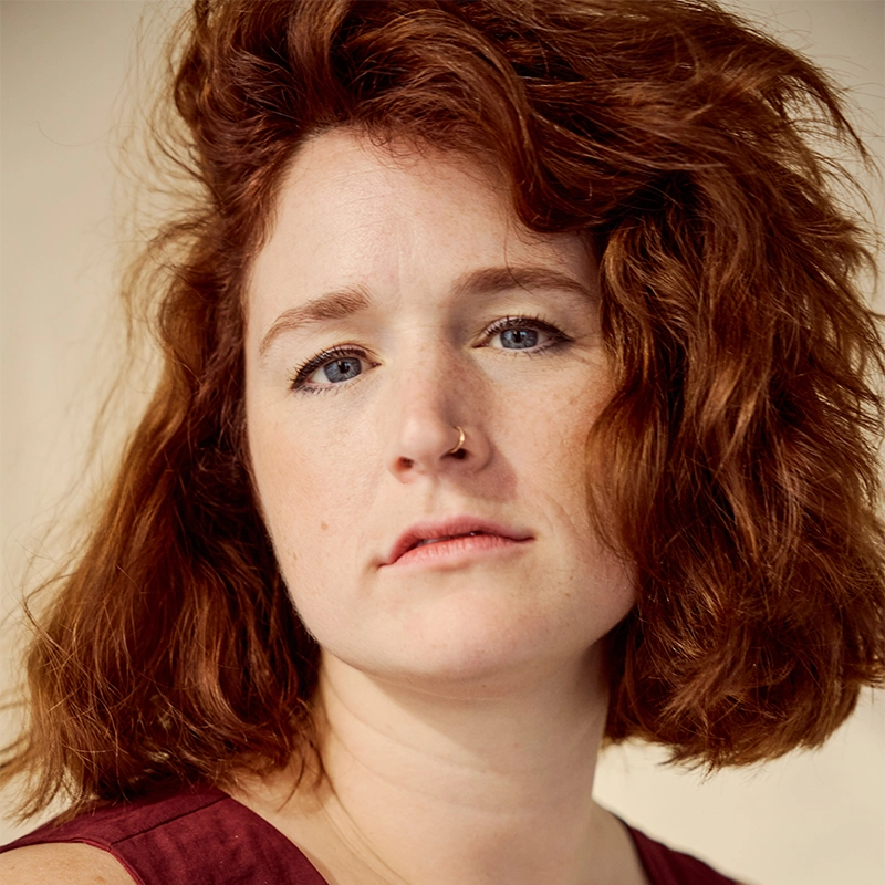 A woman with fair skin and wavy, shoulder-length red hair looks at the camera. She has blue eyes, a nose ring in her left nostril, light freckles, and a serious expression—captured in Aaron Valentine’s warm, soft lighting.