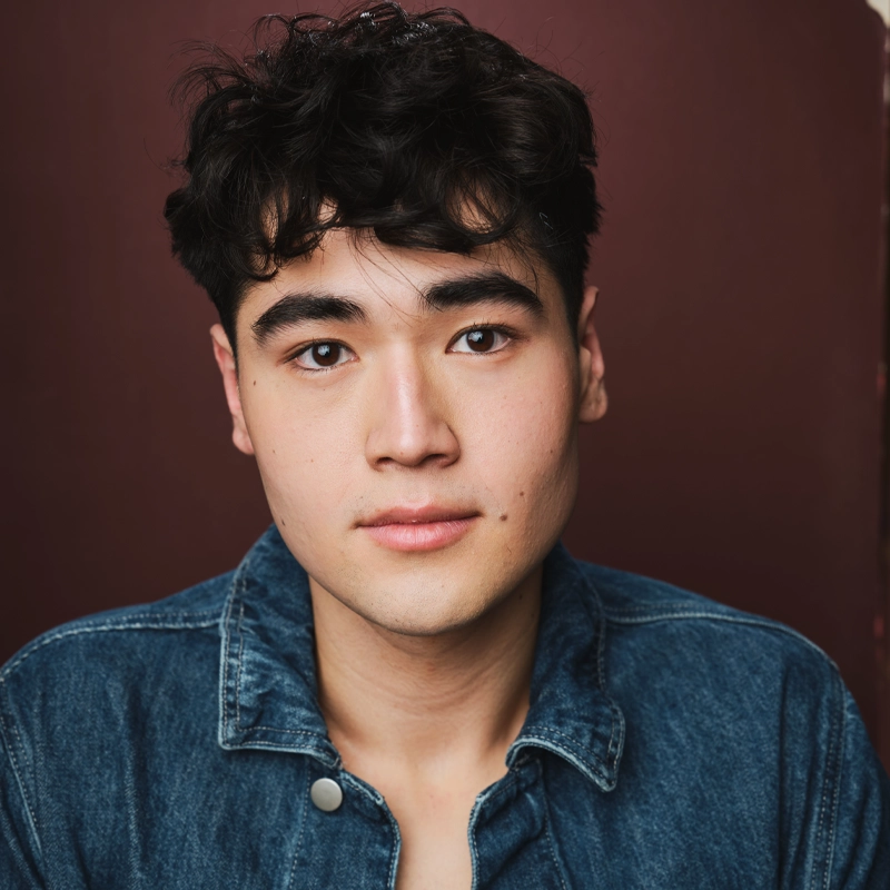 A young man, Ethan Jih-Cook, with short, wavy dark hair, fair skin, and brown eyes looks at the camera. He wears a blue denim jacket over a bare chest. The background is a plain, deep reddish-brown color. His expression is calm and neutral.